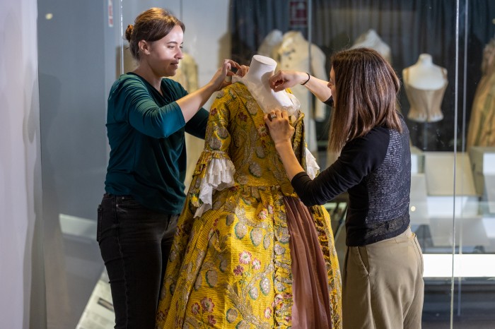 Textile Conservator Sarah Glenn and Collection Manager Elly Summers taking a 1760s yellow and gold woven silk robe à la française off display in the Fashion Museum galleries
