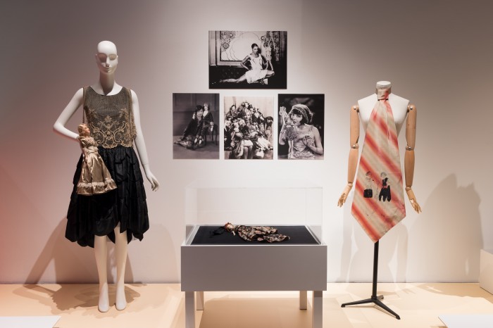 “Flappers and Boudoir Dolls” featuring a 1920s dress and doll (left) and oversize silk tie printed with an Erté illustration (right). In case: 1920s doll-shaped bag. Artwork on wall: 1920s actresses and entertainers posing with dolls. © Museum at FIT