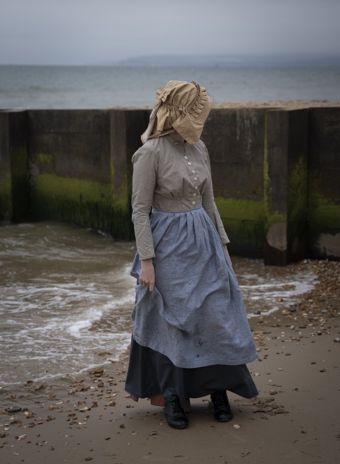 Fishergirl at the sea’s edge. Image of late 19th century working woman’s outfit. Model: Sophie Bowyer. Photographer: Kheyal Roy-Meighoo. 